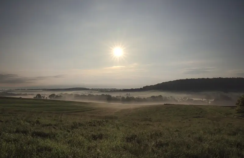 Fotografia przedstawia pagórkowaty krajobraz o świcie. Wschodzi słońce, mgły ścielą się w dolinach. Niebieskie niebo i zieleń pól.