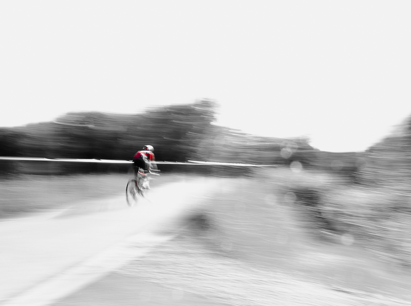 Rowerzysta w ruchu. Kolor ubrania czerwony, otoczenie czarno-białe.