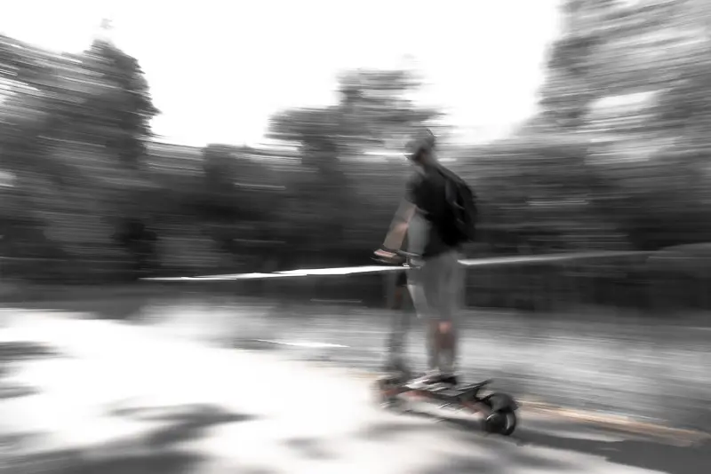 Fotografia przedstawia mężczyznę, a może chłopca na hulajnodze. Postać uchwycona w ruchu, rozmazane tło.